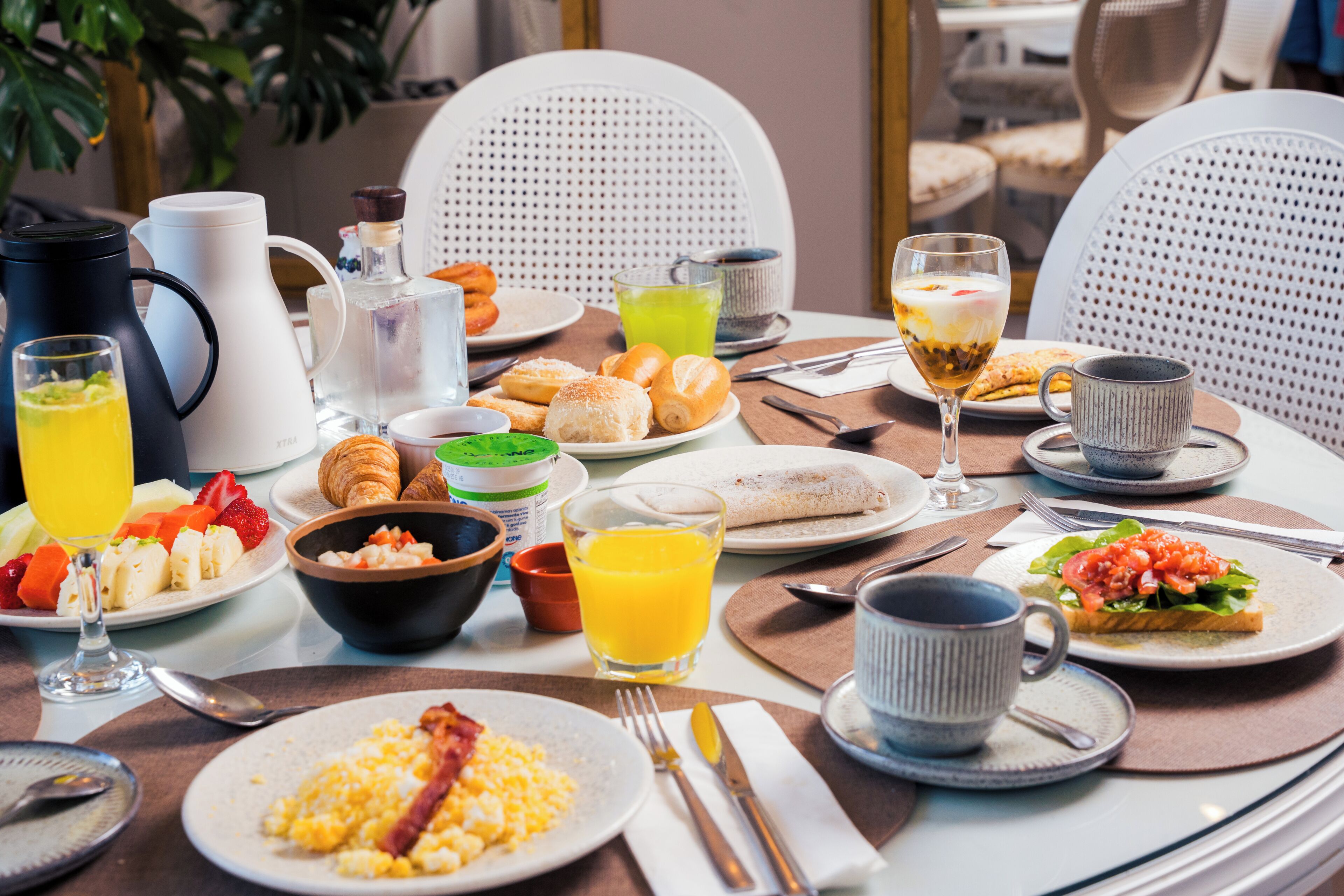 Un desayuno con diferentes alimentos, una cafetera y un vaso de zumo de naranja sobre una mesa redonda.