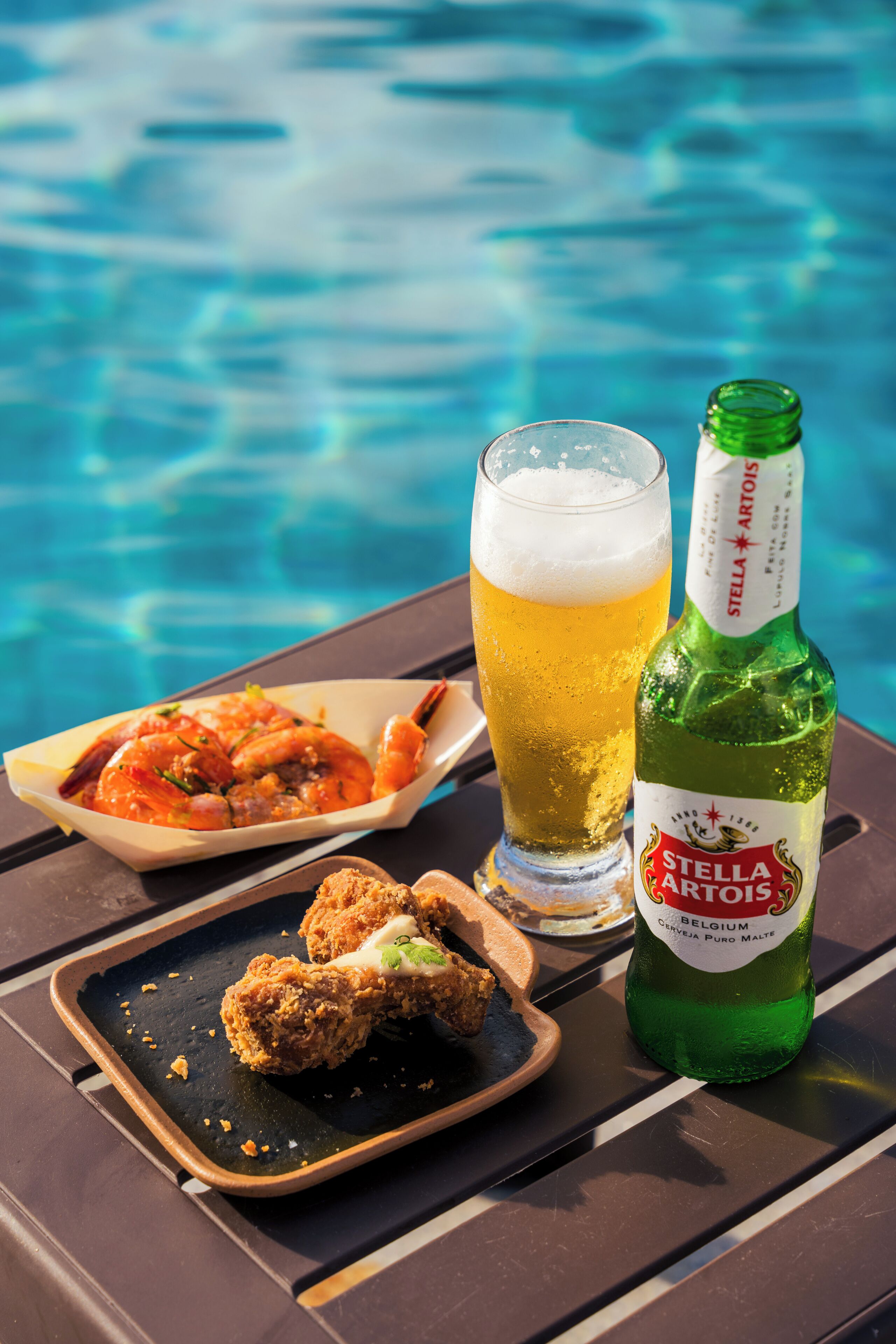 A table with a bottle of Stella Artois beer, a glass of beer, and a plate of food by a pool.