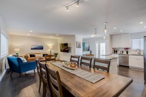 Eine moderne Küche mit weißen Schränken, ein Essbereich mit einem Holztisch und -stühlen sowie ein Wohnzimmer mit einem blauen Sofa und einem an der Wand befestigten Fernseher.