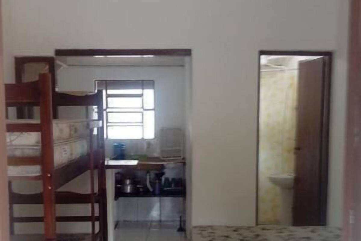 Um quarto com uma cama de solteiro, uma escrivaninha com cadeira, uma janela, um ventilador de teto e uma porta.