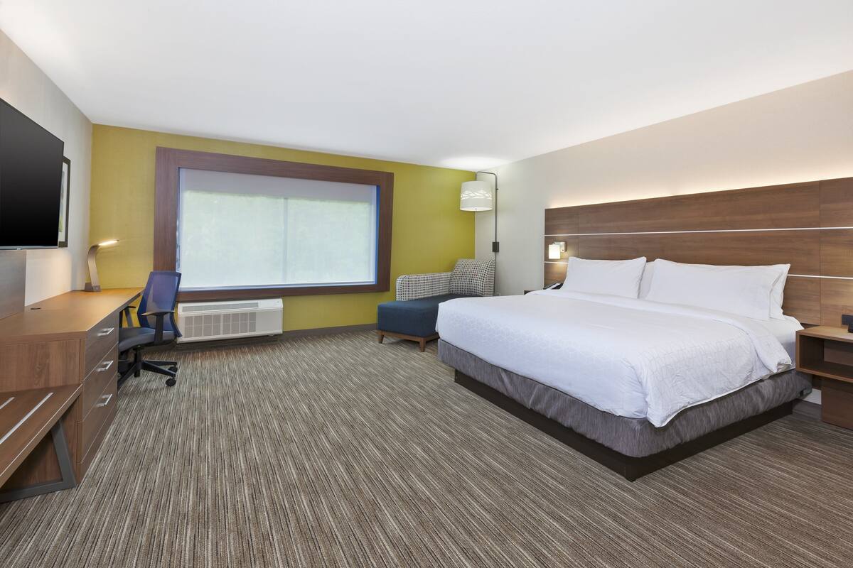 A hotel room with a large bed, a desk, a chair, a window, and a wall-mounted TV.