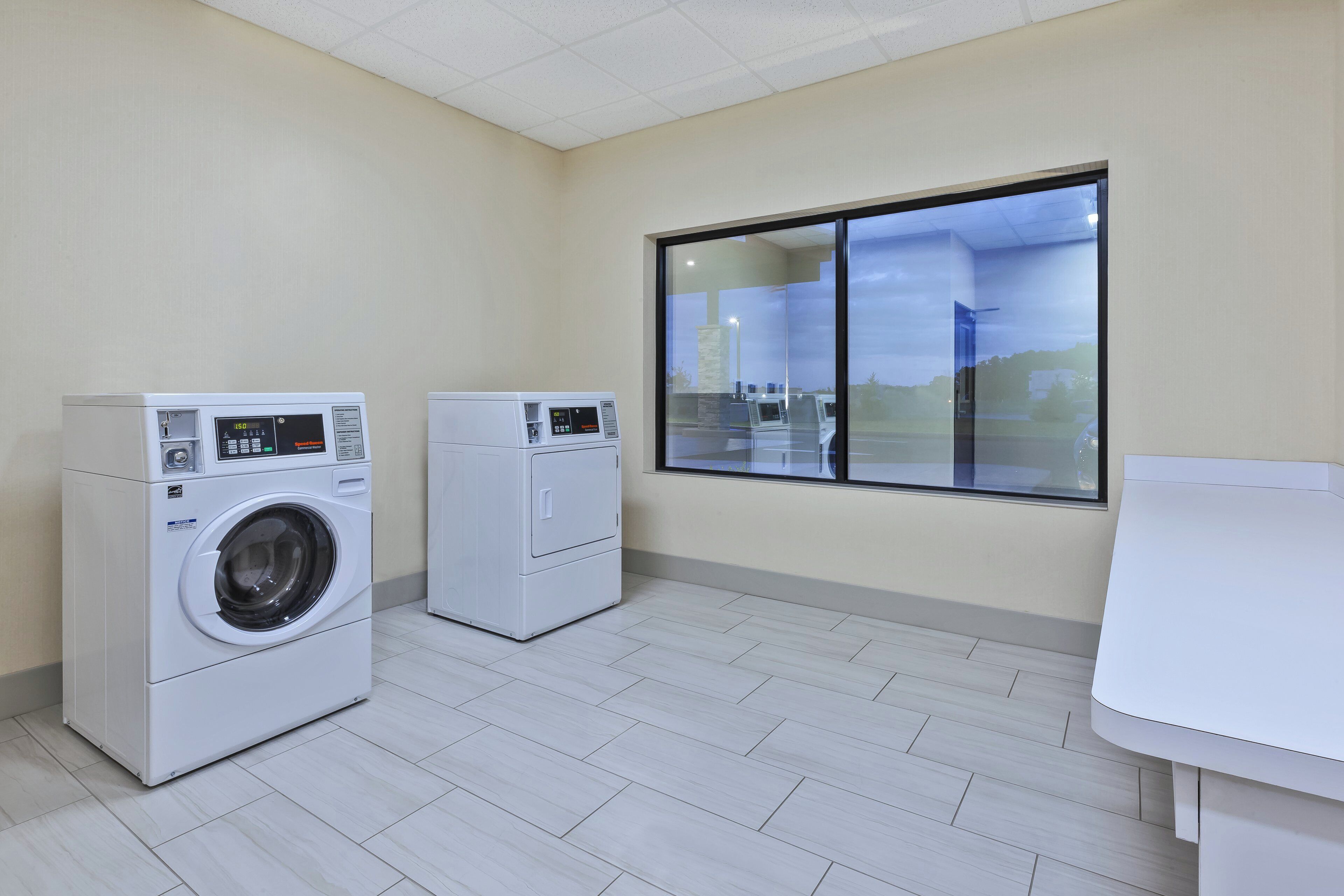 Een wasserette met twee wasmachines, een raam met uitzicht op een gebouw en een betegelde vloer.