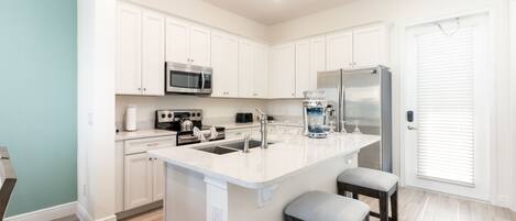 A modern kitchen with white cabinets, stainless steel appliances, a central island with stools, and a large window with blinds.