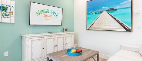 A room with a teal wall, a white cabinet, a wooden coffee table with a tray, and a large framed picture of a beach scene.