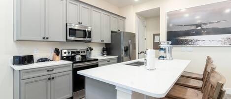 A modern kitchen with grey cabinets, stainless steel appliances, a white countertop, and a wooden dining area.