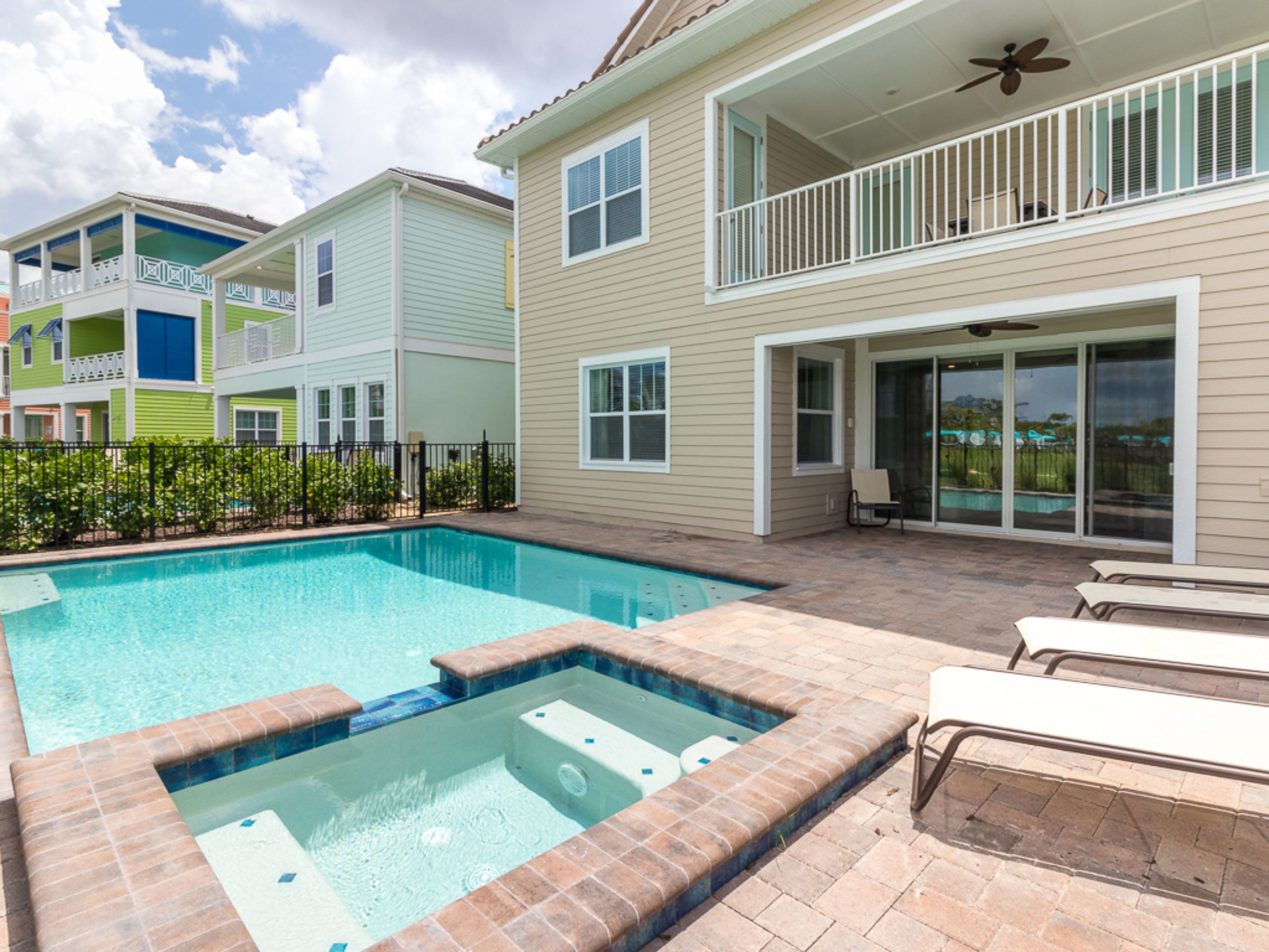 A residential swimming pool with a hot tub, surrounded by a patio with chairs and a covered deck area.