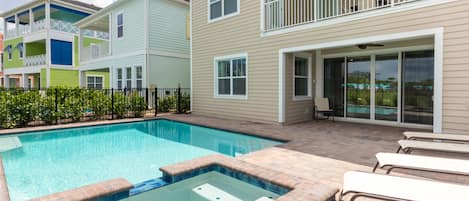 A residential swimming pool with a hot tub, surrounded by a patio with chairs and a covered deck area.