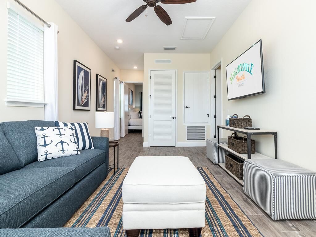 Un salon moderne avec un canapé gris, un pouf blanc et un tapis à rayures. On y trouve un ventilateur de plafond, un tableau encadré au mur et une petite table avec des paniers.