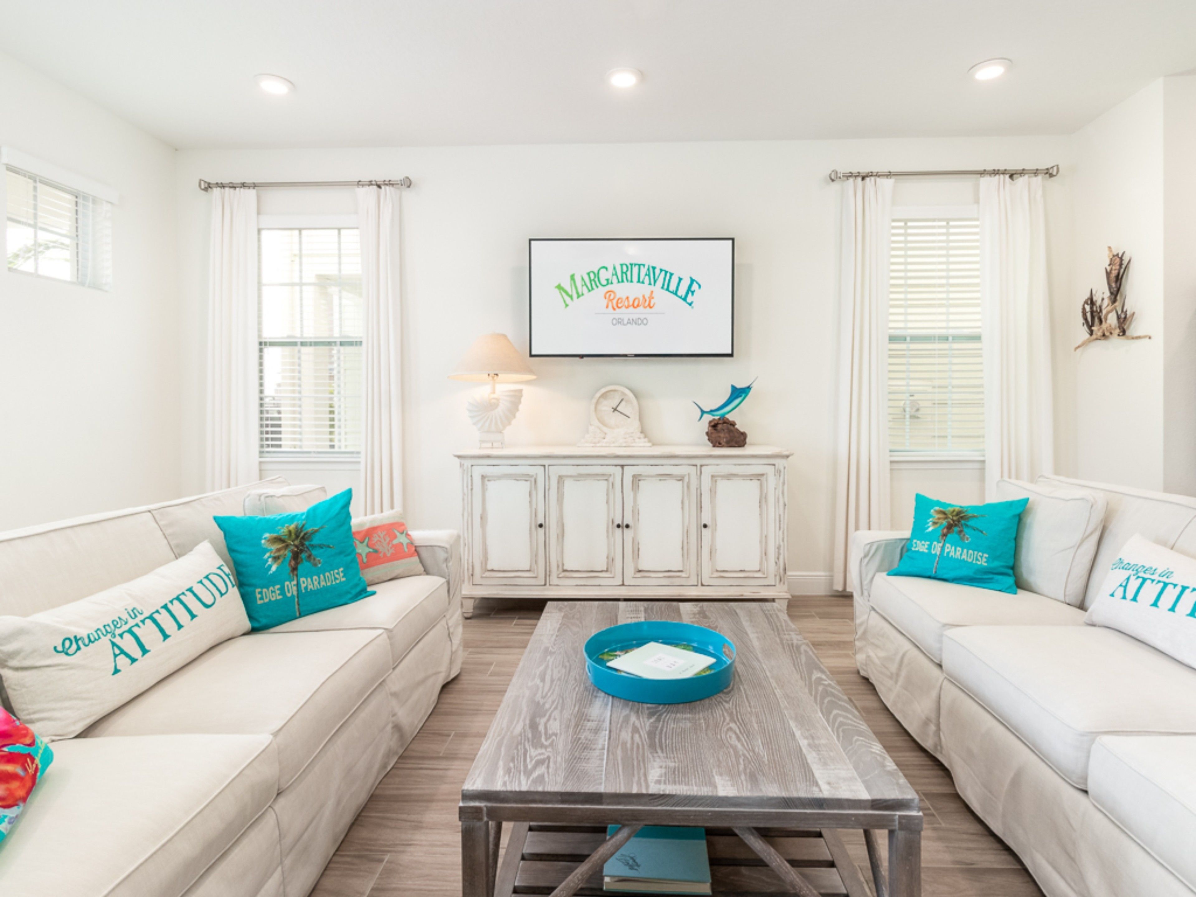 A bright living room with two white sofas, each with teal pillows, a wooden coffee table, and a white cabinet with decorative items. A framed sign on the wall reads "Marriott Village Resort Orlando".