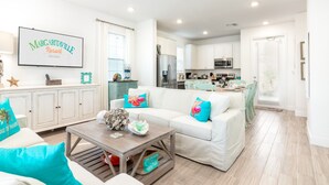 Un salon spacieux avec un canapé blanc, des coussins turquoise, une table basse et une cuisine en arrière-plan.
