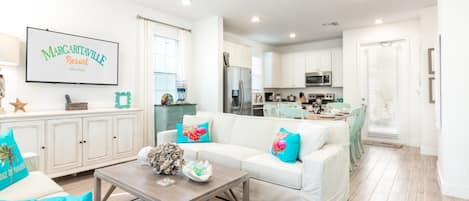 A spacious living room with a white sofa, teal cushions, a coffee table, and a kitchen in the background.