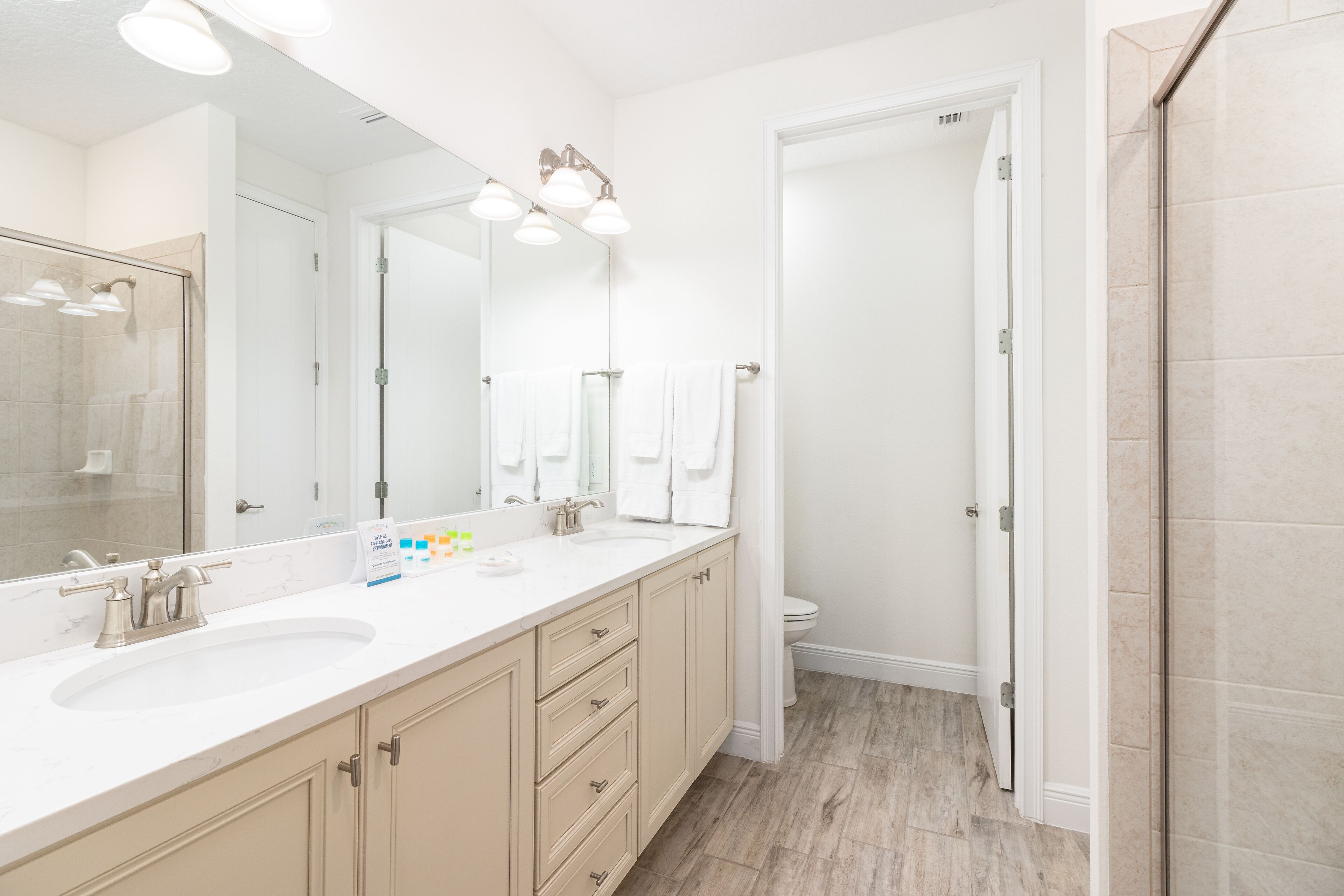 A modern bathroom with a large mirror, two sinks, a shower, and a toilet.