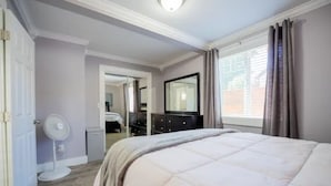 Et soveværelse med seng, kommode, spejl, ventilator og et vindue med gardiner.