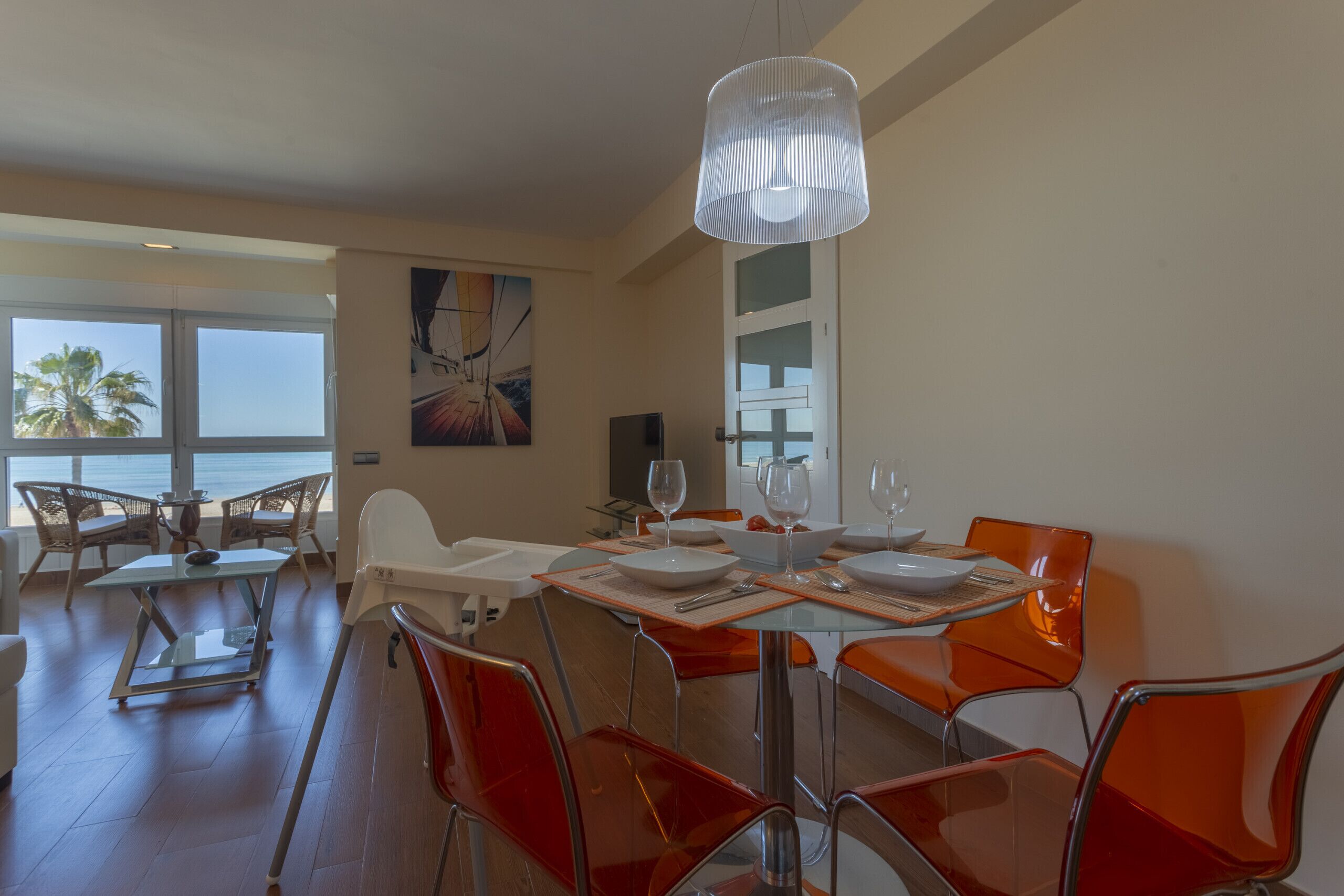 Un comedor con mesa de vidrio, sillas naranjas y una lámpara colgante blanca.