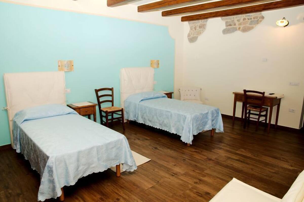 A room with two single beds, wooden chairs, a small table, and a stone wall.