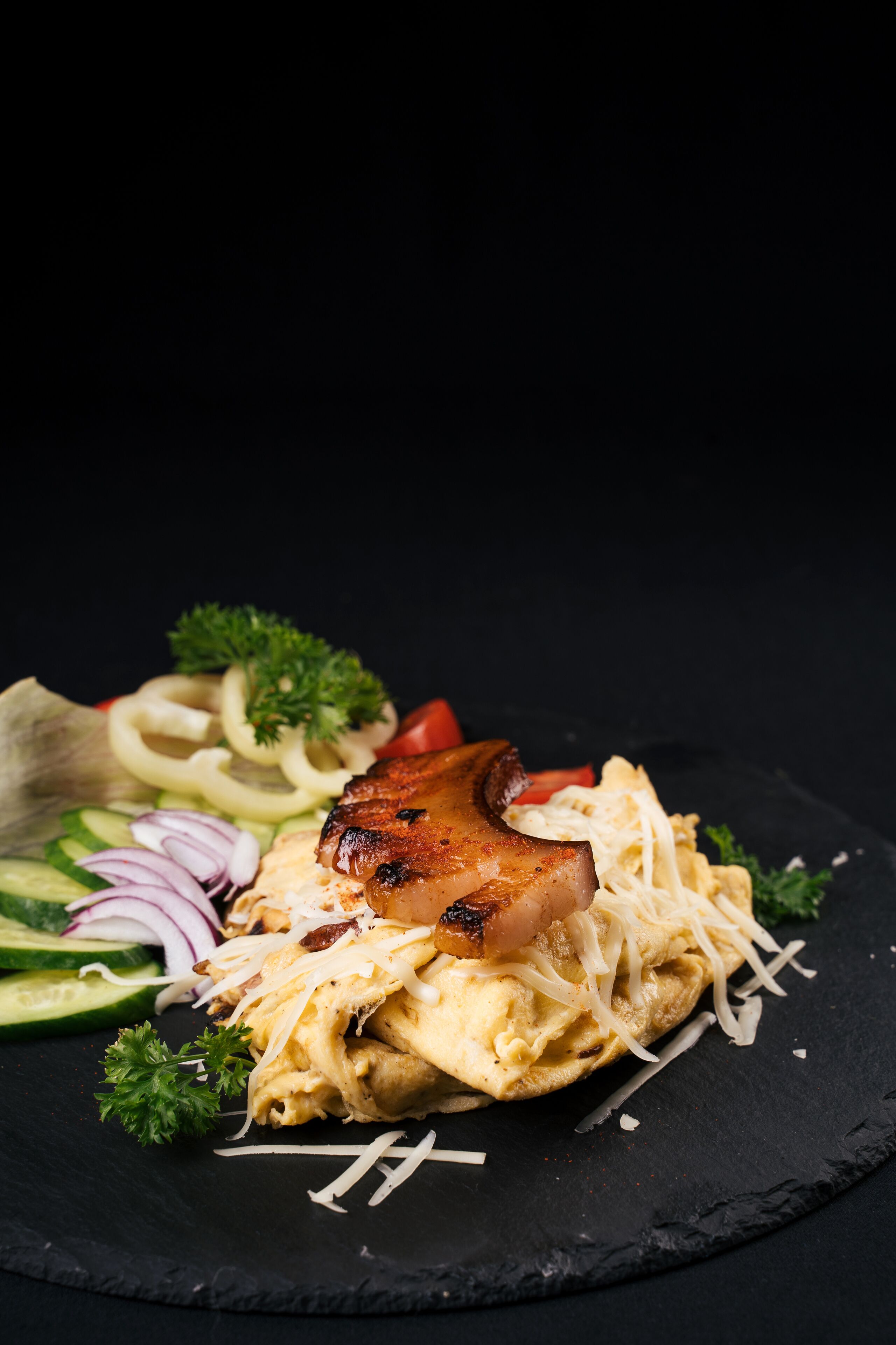 A black slate plate with a grilled meat, shredded cheese, and a salad including cucumbers, red onions, and parsley.