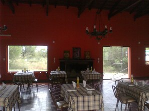 A dining area with tables set for two, a fireplace, and large windows offering a view of the outdoors. - Hotel Colonial (Merlo)