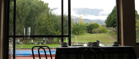 Un comedor con una mesa y sillas, vista a una piscina y un jardín con árboles y montañas al fondo.