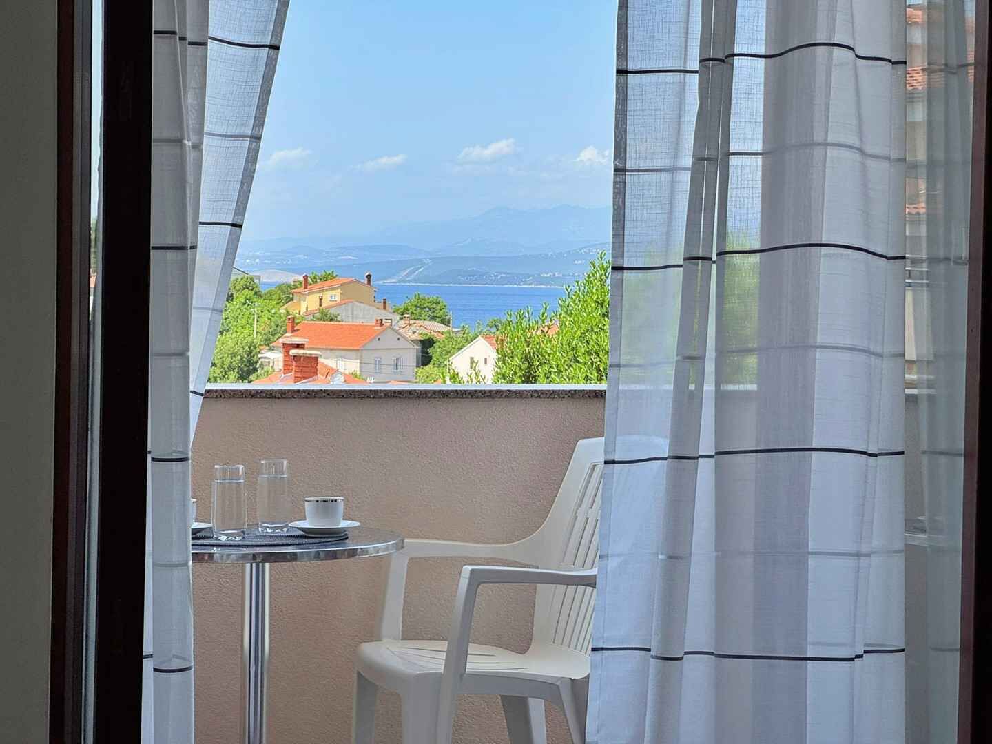 Ein Balkon mit Tisch und Stuhl, mit Blick auf eine Küstenstadt mit Häusern und Meerblick.