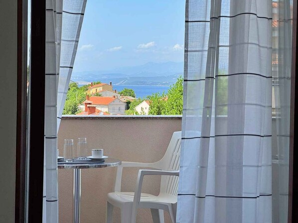 A balcony with a table and a chair, overlooking a coastal town with houses and a view of the sea.