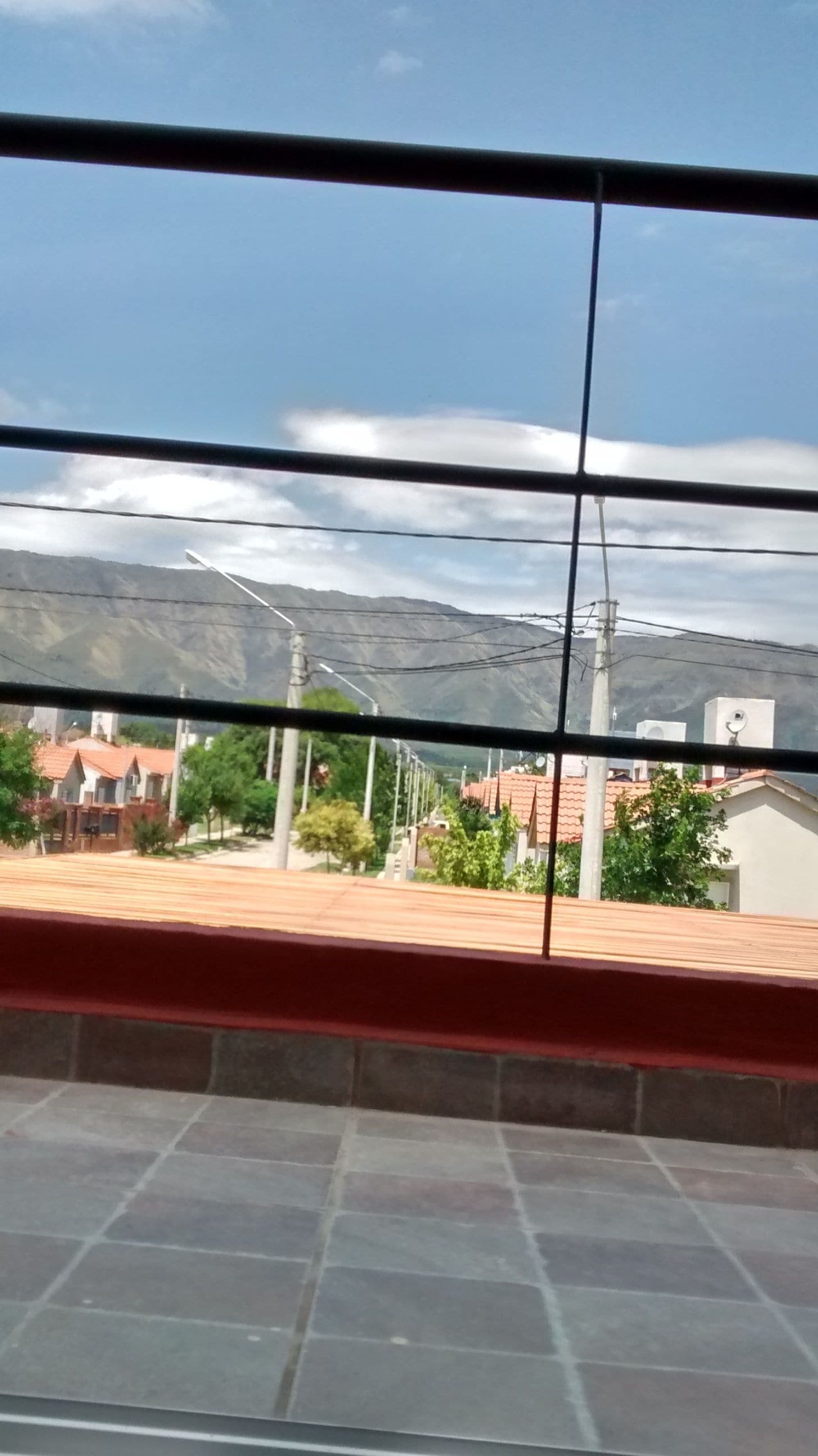 Blick durch ein Fenster mit Metallgitter, der einen Wohnbereich mit Häusern, Bäumen und einem Berg im Hintergrund zeigt.
