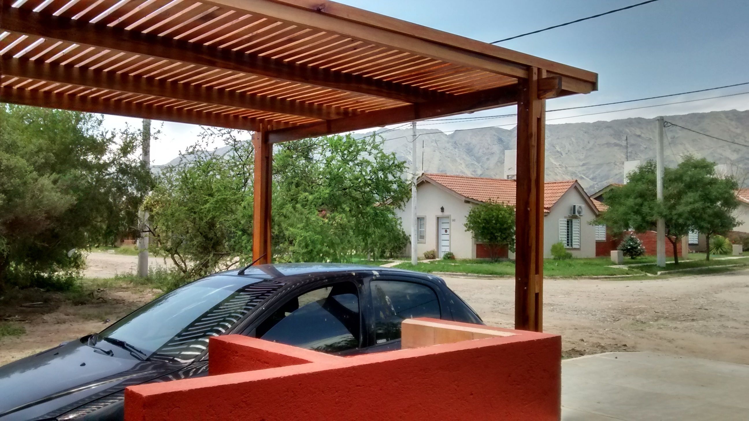 A car parked under a wooden pergola with a view of houses and mountains in the background.