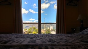 A bed with a patterned bedspread, a window with curtains, and a view of buildings and mountains.