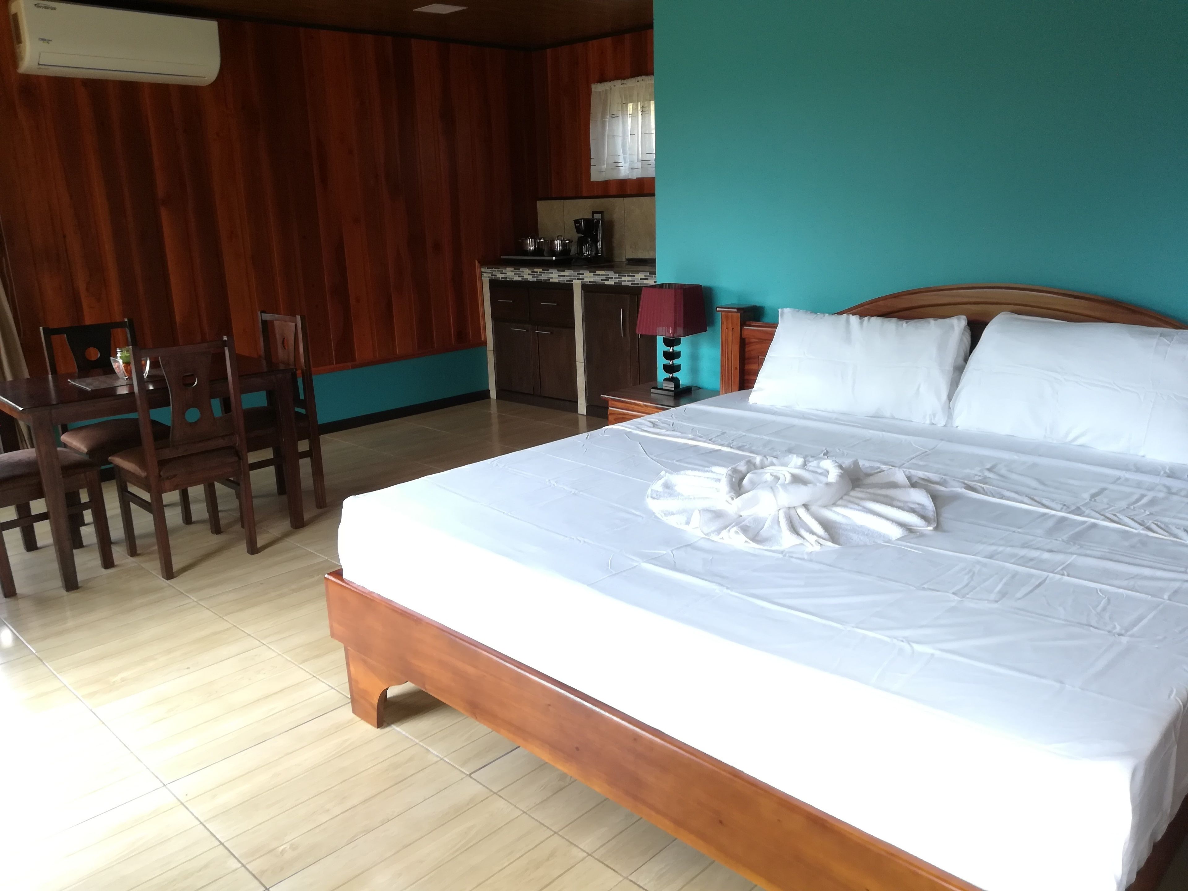 A hotel room with a wooden bed, a small kitchenette, and a dining area with chairs.