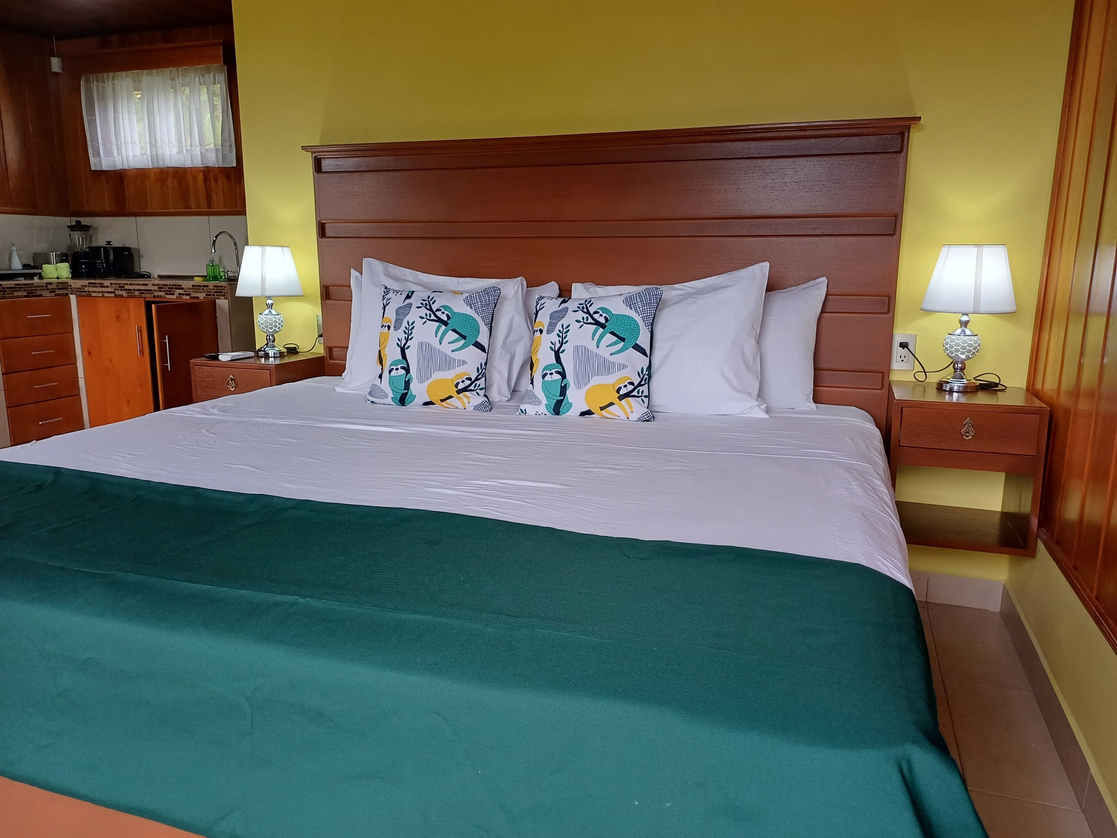 A hotel room with a large bed, two bedside tables with lamps, a wooden headboard, a green throw blanket, and a kitchen area in the background.