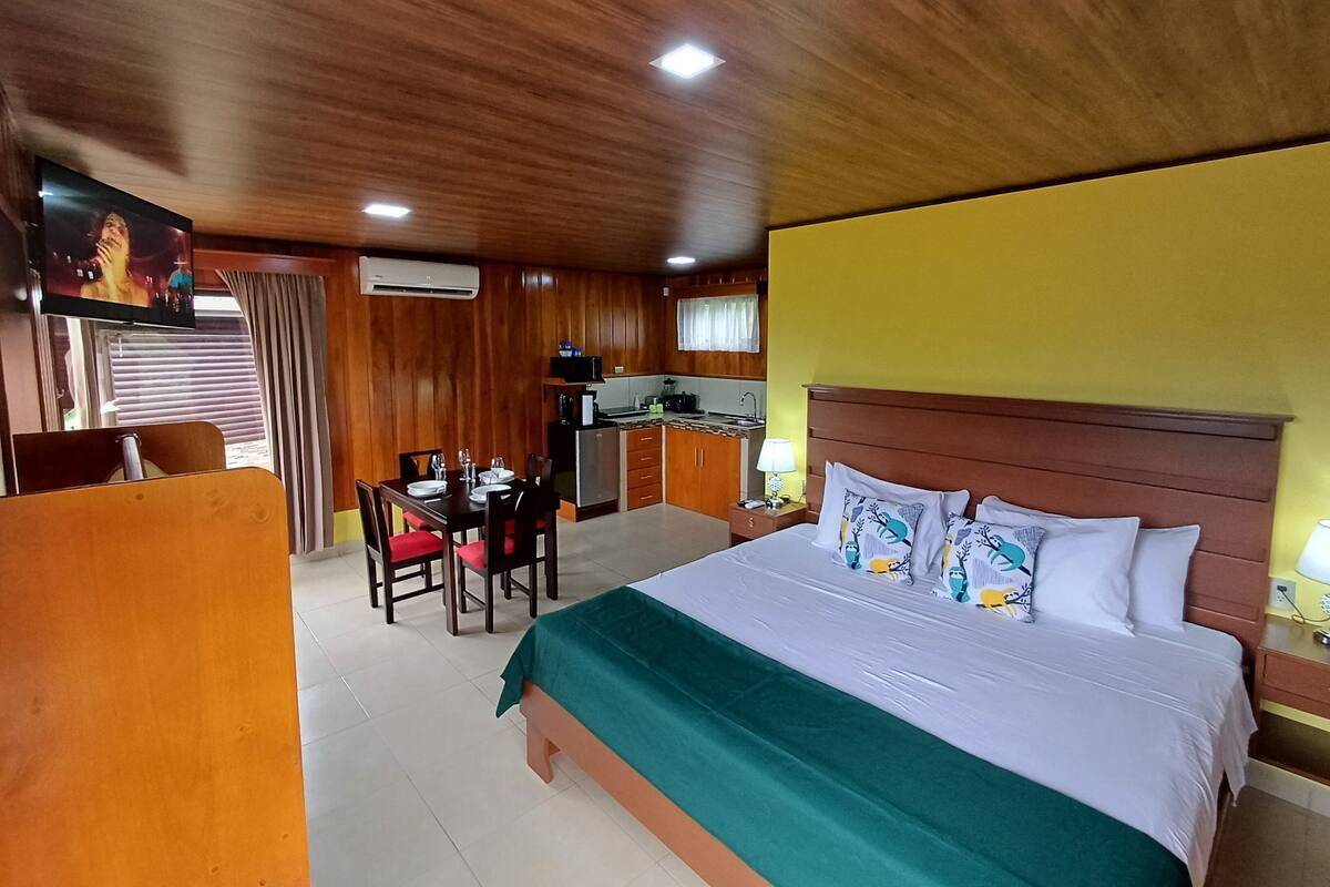 A hotel room with a wooden ceiling, a bed with a green bedspread, a dining table with chairs, a kitchenette, and a television on the wall.
