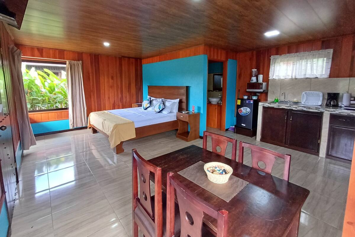 A room with a bed, a dining table with chairs, a kitchen area, and a view of greenery outside.