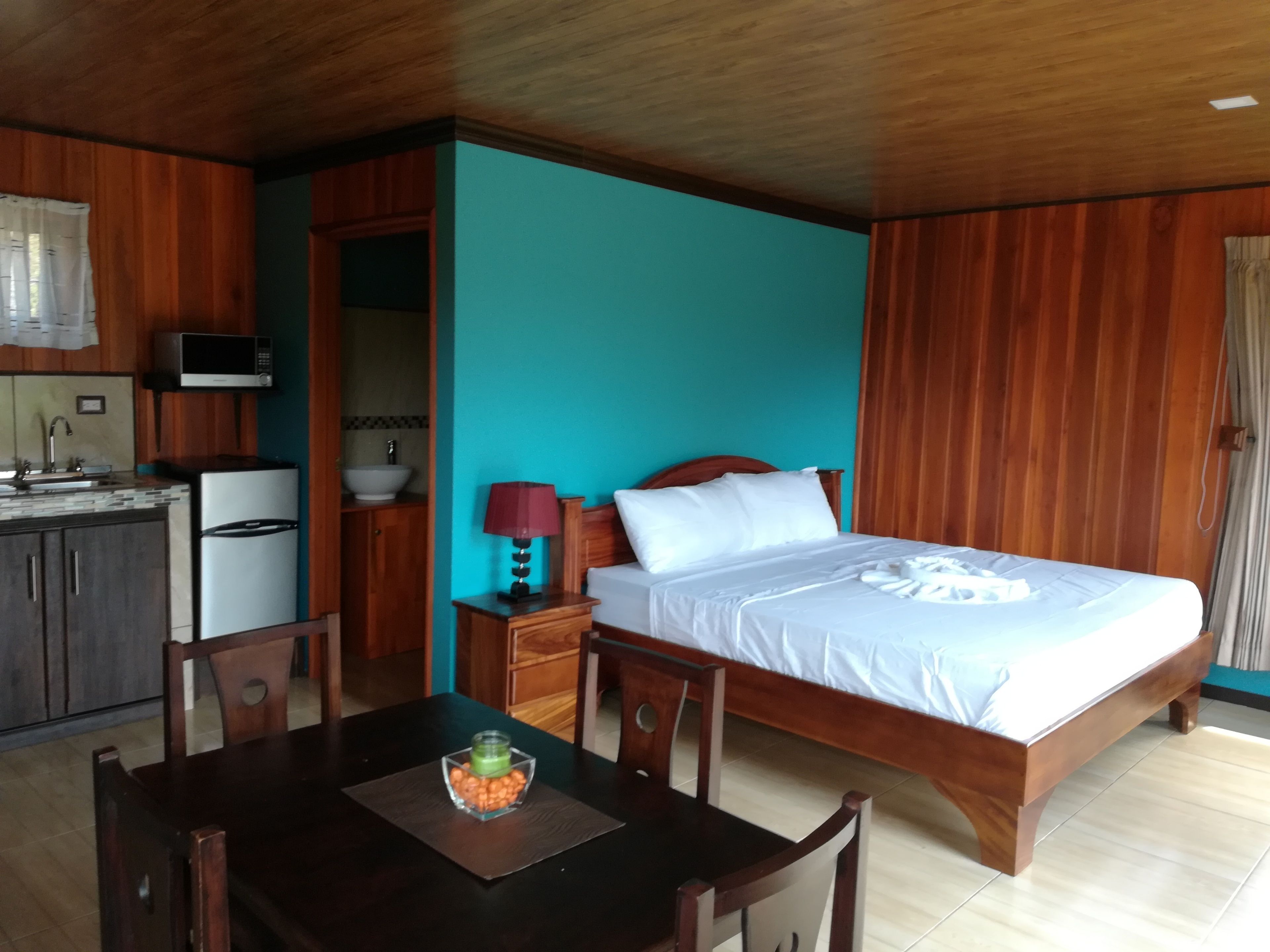 A hotel room with a wooden bed, a small bedside table with a lamp, a wooden dining table with chairs, a kitchen area with a microwave, sink, and refrigerator.