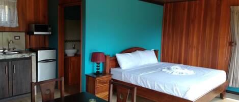 A hotel room with a wooden bed, a small bedside table with a lamp, a wooden dining table with chairs, a kitchen area with a microwave, sink, and refrigerator.