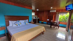 Quarto de hotel com cama, escrivaninha, cadeira, TV e janela com vista para área verde.