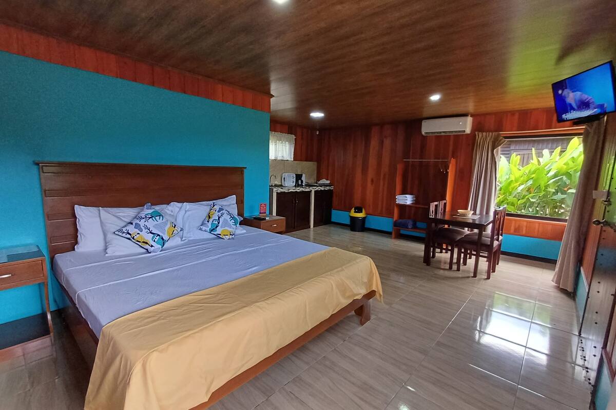 A hotel room with a bed, a desk, a chair, a TV, and a window with a view of greenery.