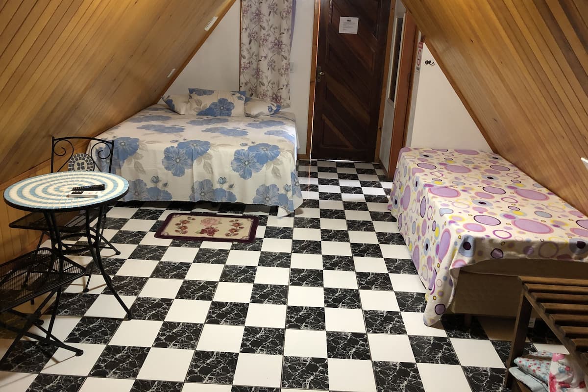 Una habitación pequeña y acogedora con piso a cuadros, una cama con ropa de cama floral, una mesita con un control remoto y una puerta con un cartel.