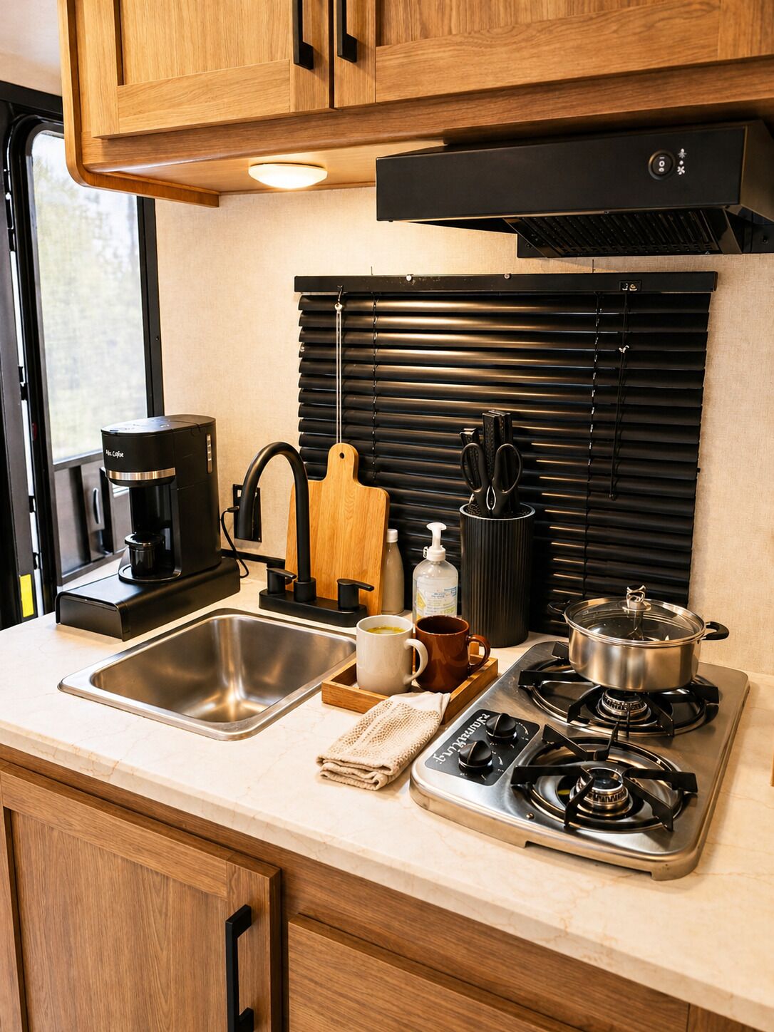 A compact kitchen with a coffee maker, a pot on the stove, a sink, and a window with blinds.