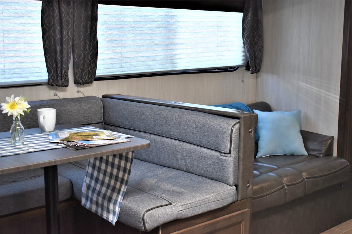 A compact RV interior with a dining area, a cushioned bench, and a window with curtains.