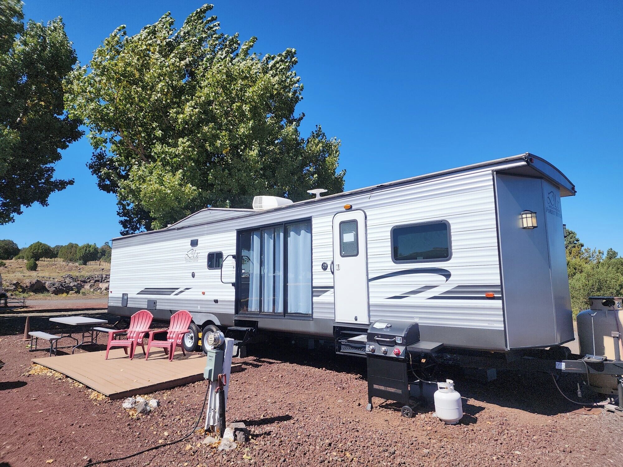 A stationary RV with a sliding door, a small attached kitchen area, and a deck with chairs.