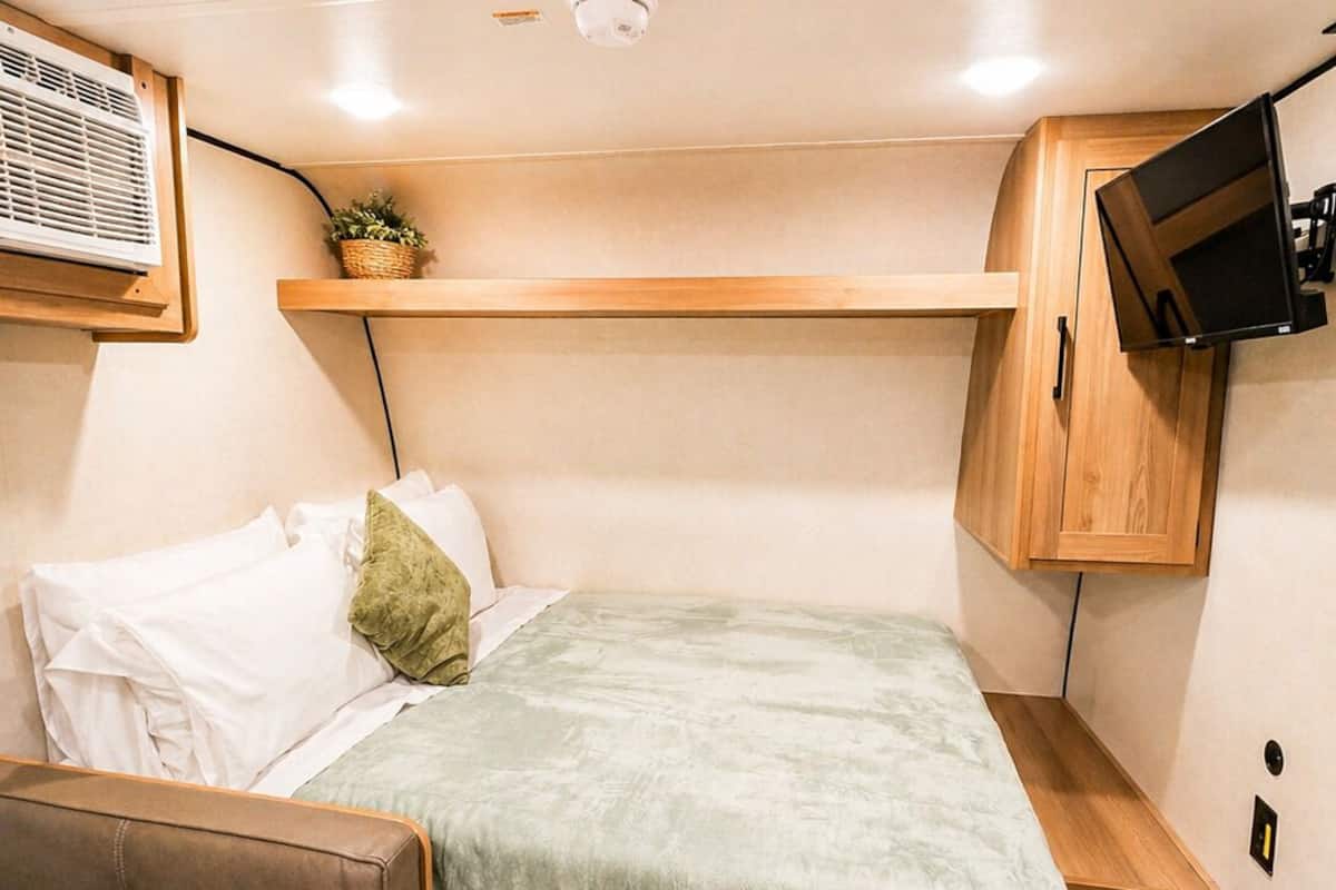 A compact RV interior with a bed, a seating area, a mounted TV, and a ceiling-mounted air conditioning unit.
