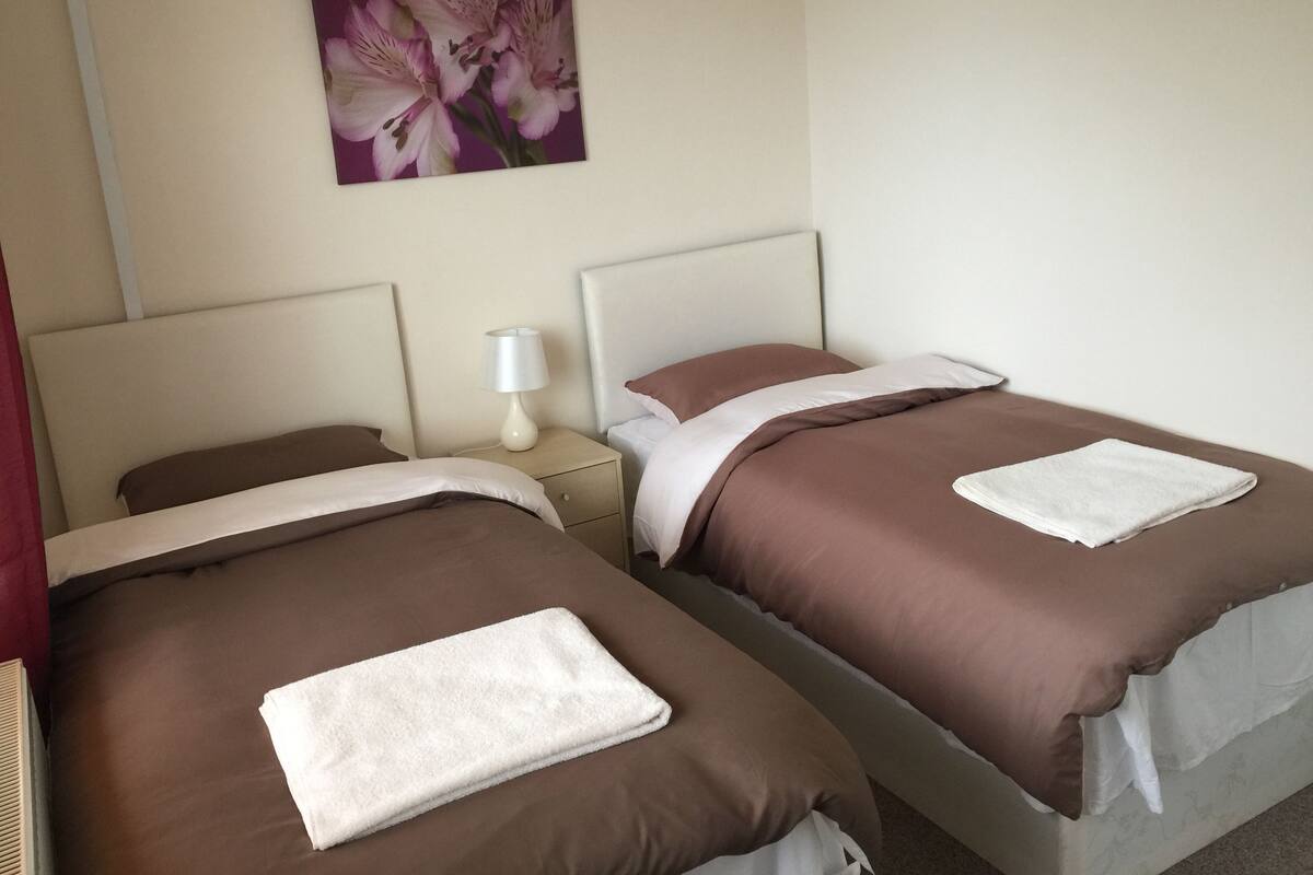 Two beds with brown bedding and white pillows, a white bedside table with a lamp, and a floral painting on the wall.