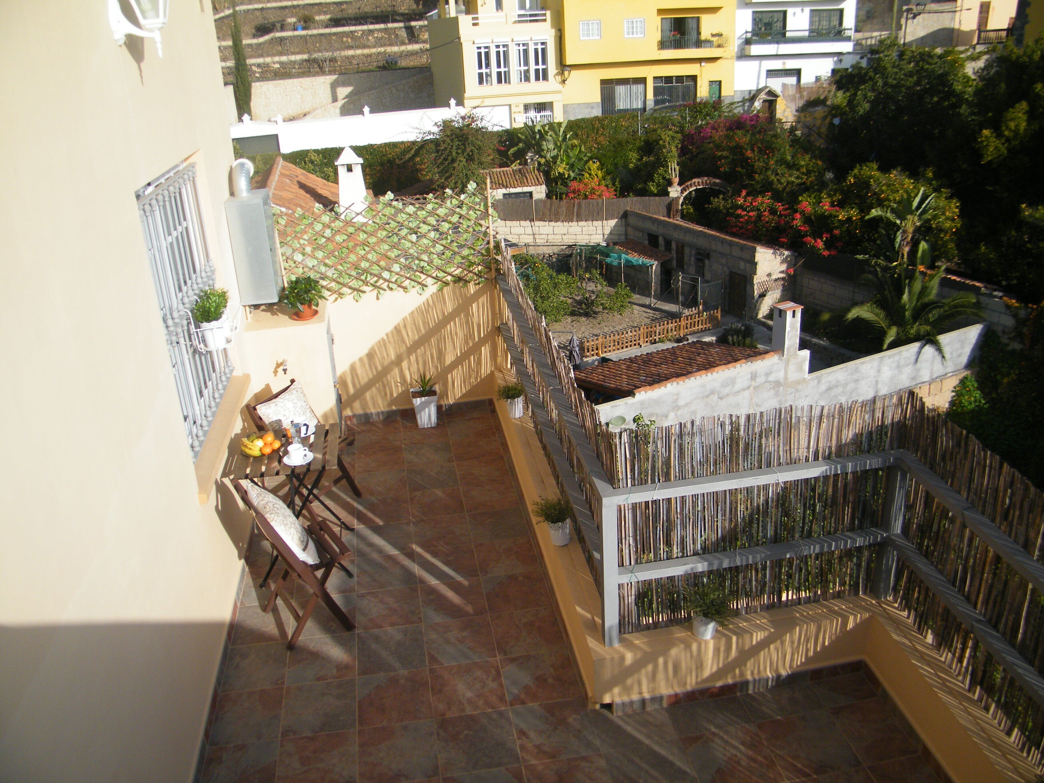 Un balcón residencial con suelo de baldosas, una silla de madera, una mesa pequeña con frutas y plantas en macetas.