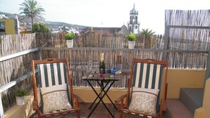 Un balcon avec deux chaises à rayures, une petite table avec une bouteille et des verres, et une vue sur une église et des bâtiments en arrière-plan.