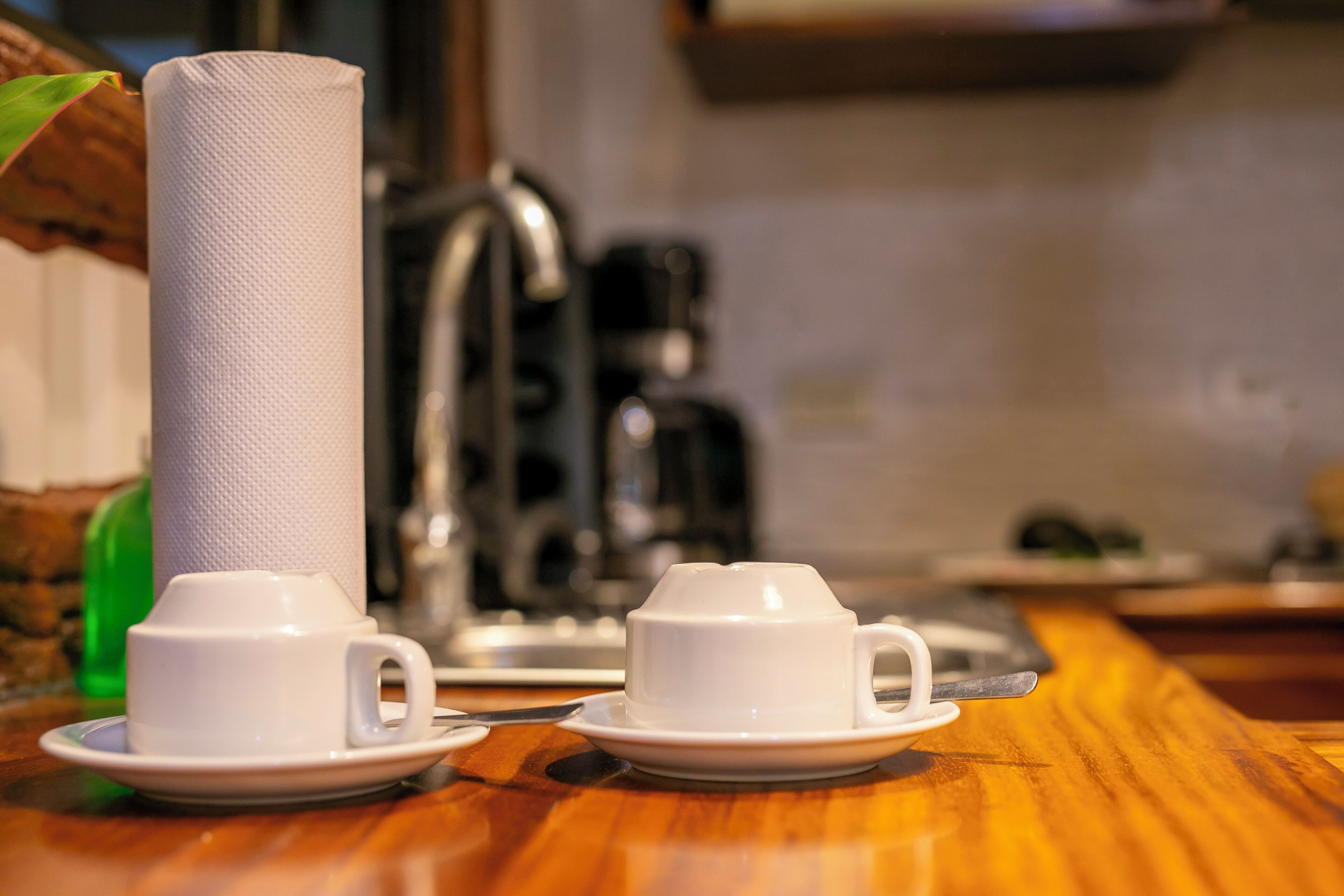 Una mesa de madera con dos tazas y platillos blancos, un rollo de papel de cocina y una cafetera.
