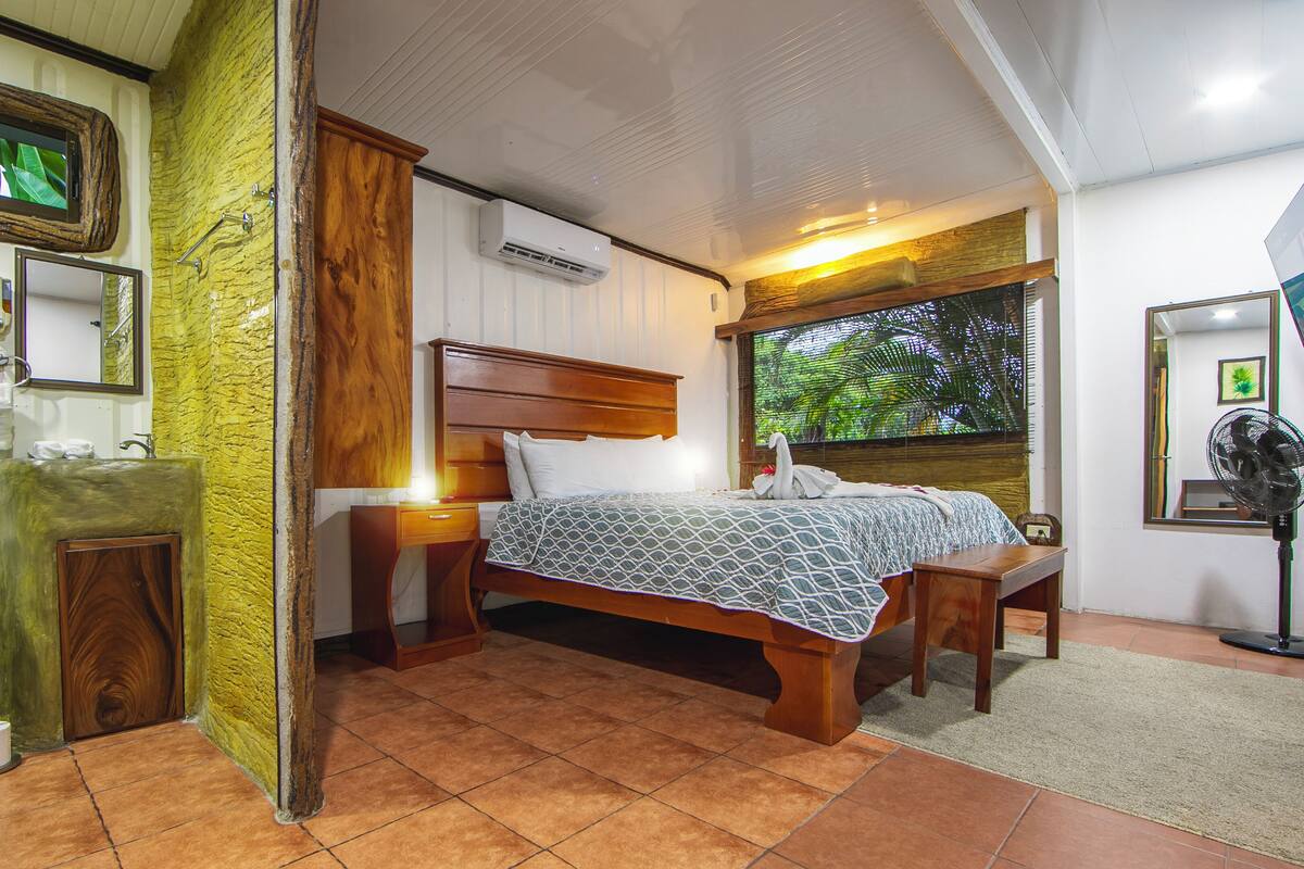 Une chambre avec un lit en bois, une table de chevet, une salle de bain avec des toilettes et un lavabo, et une grande fenêtre donnant sur un paysage verdoyant.