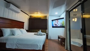Quarto com cama, televisão fixada na parede, um banco e porta de vidro corrediça para um banheiro.