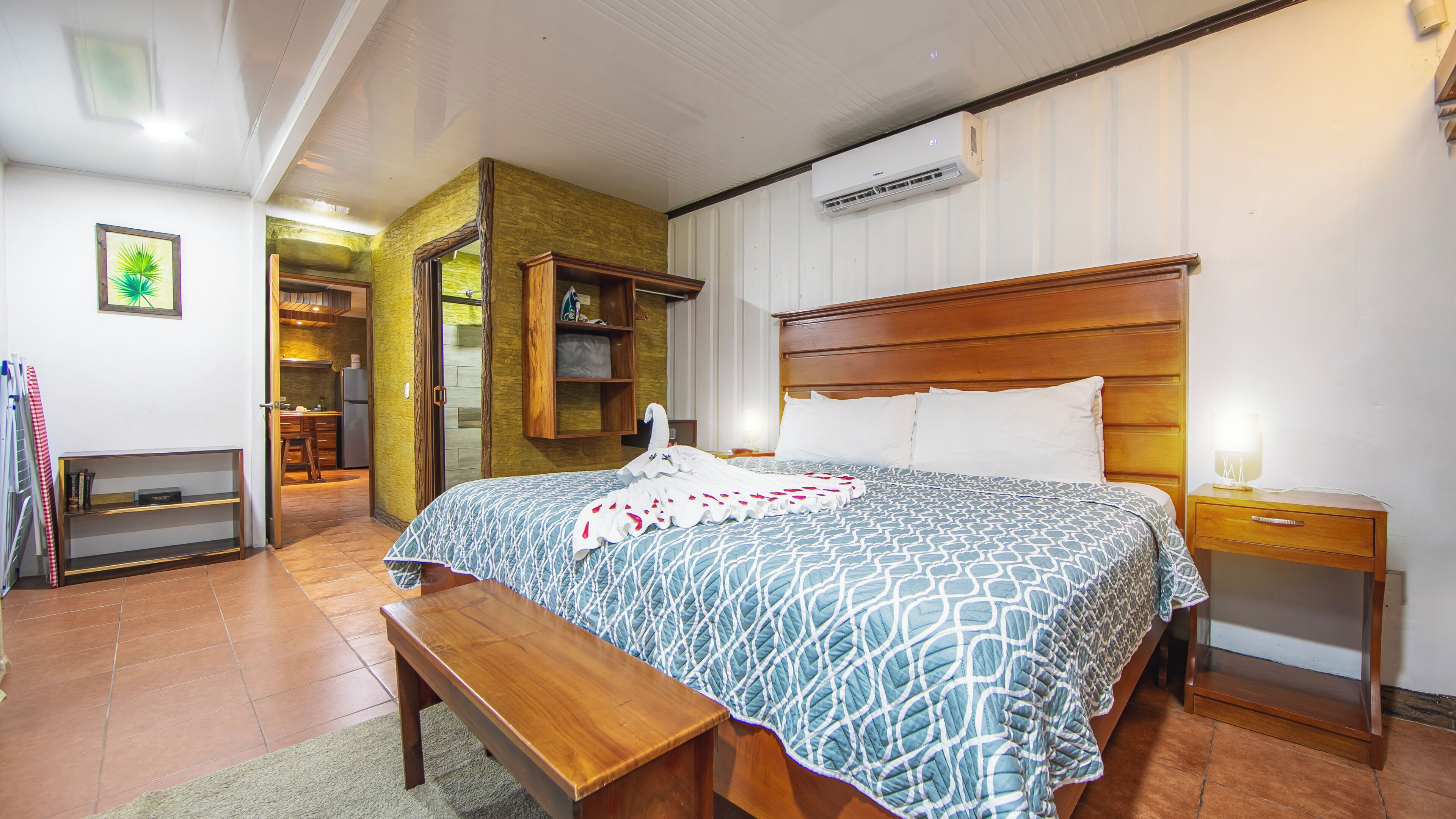 Habitación de hotel con una cama de madera, un banco, una mesita de noche con lámpara, un armario y un equipo de aire acondicionado.