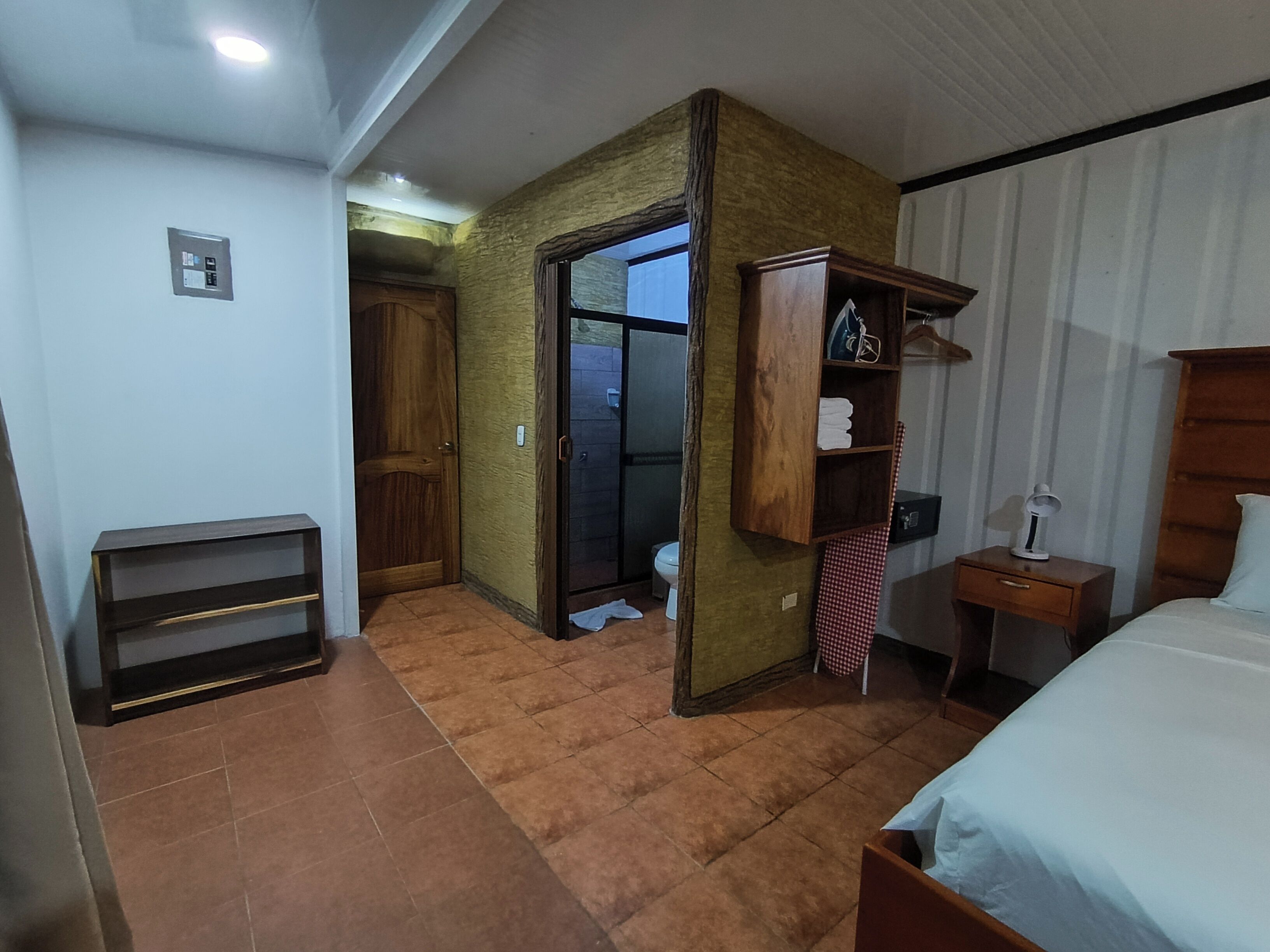 Un dormitorio con cama, armario de madera, mesita de noche con lámpara y puerta que da a un baño.