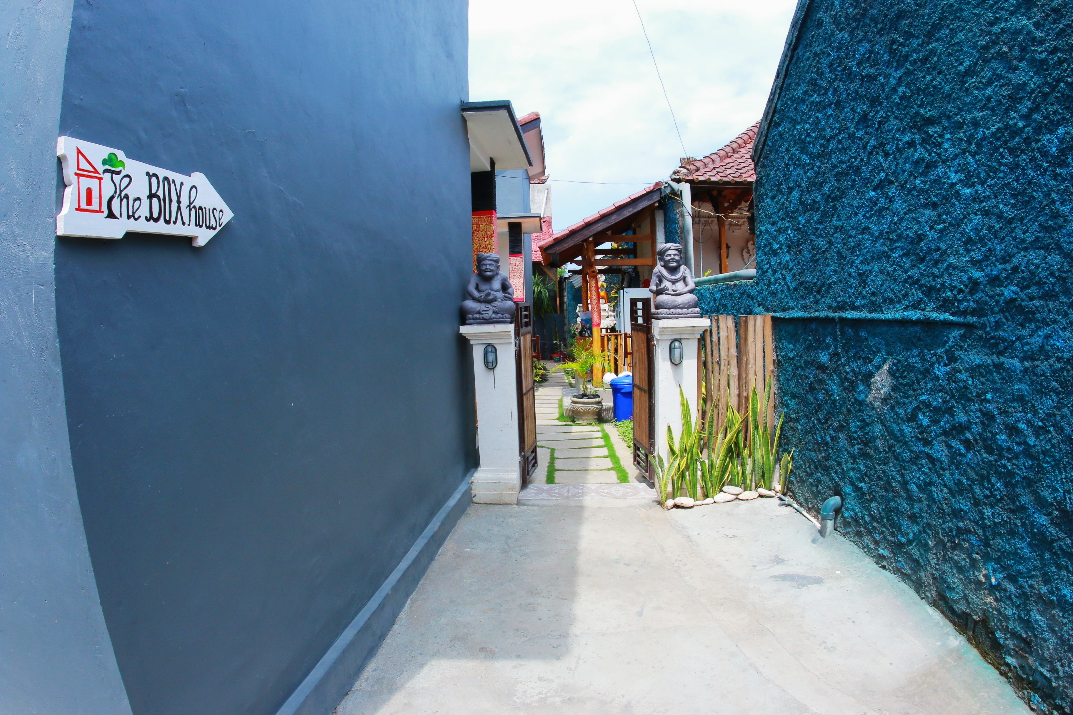 A narrow alleyway with a sign for 'The BOX house' on the left, leading to a wooden structure with a red roof.