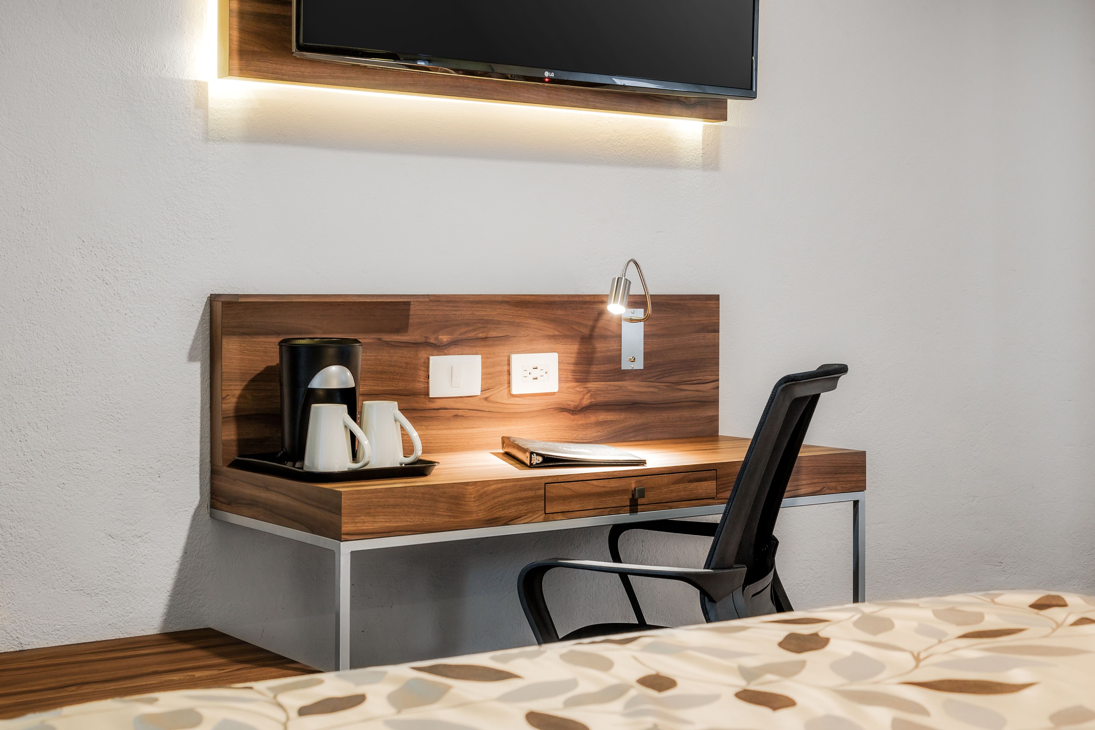 A modern hotel room with a wooden desk, a black chair, a coffee maker, and a flat-screen TV mounted above the desk.